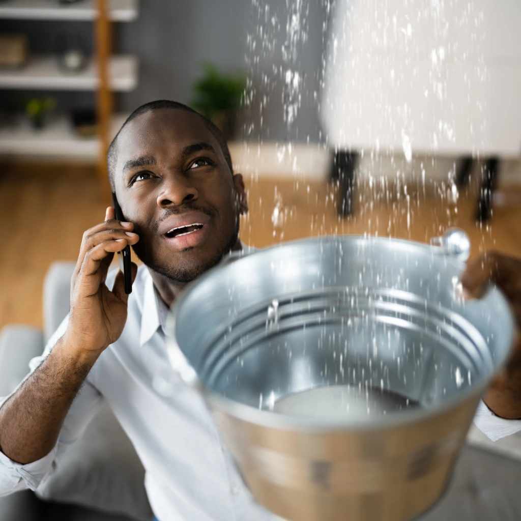 Water leaking from the ceiling and the man catching the water in a bucket while calling a plumber